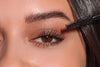 Frau mit geschminkten Augen, die eine Mascara Bürste vor ihr Gesicht vorhält