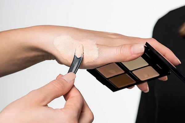 Es wird eine Hand gezeigt die eine befüllte Palette in der Hand hält und sich mit einer Spatel Camouflage auf die Hand aufträgt