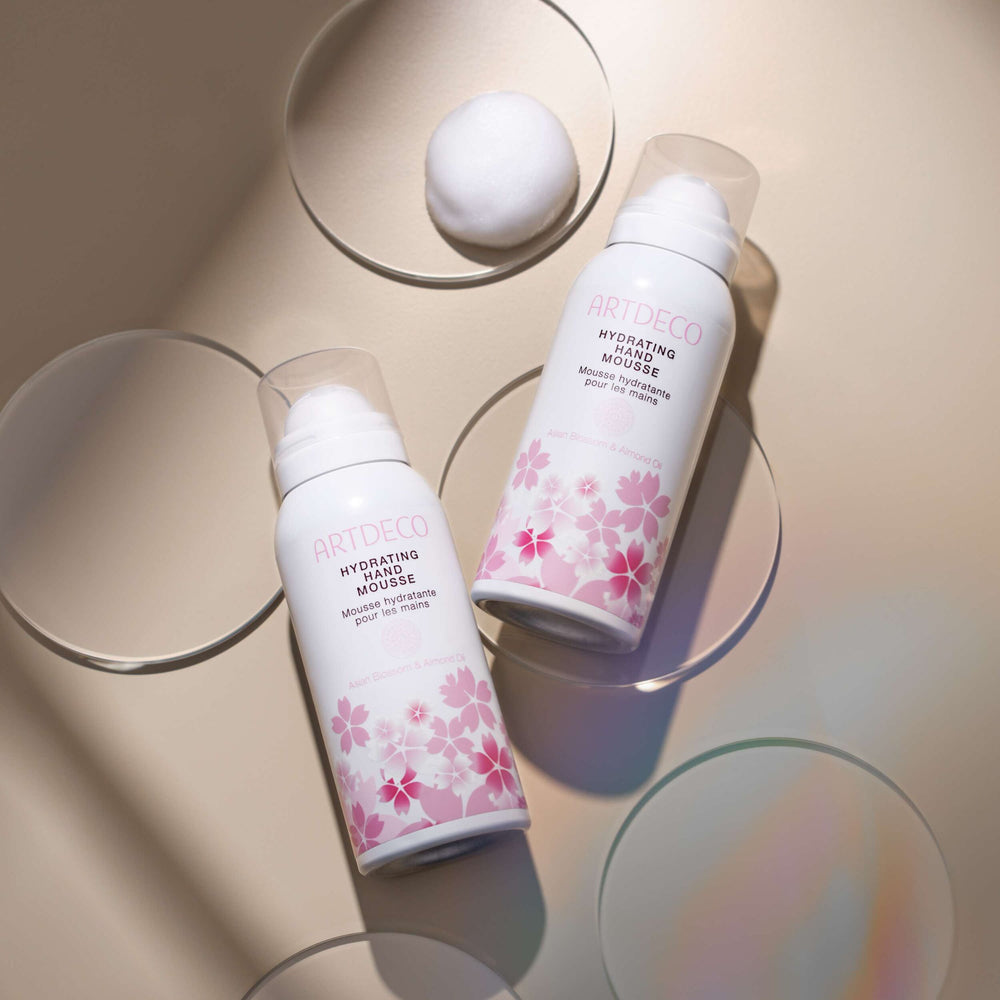 Zwei Hydrating Hand Mousse liegen auf einem neutralen Hintergrund. Neben einer der Flaschen kann an die Mousse-Textur des Produktes sehen