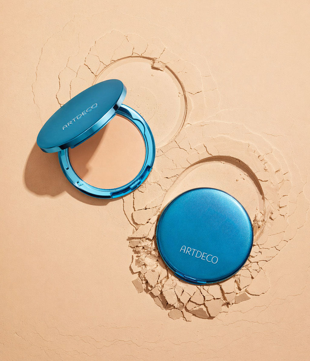 Eine geschlossene und eine geöffnete Sun Protection Powder Foundation liegen auf einem Untergrund aus Puder, der wirkt wie Sand.