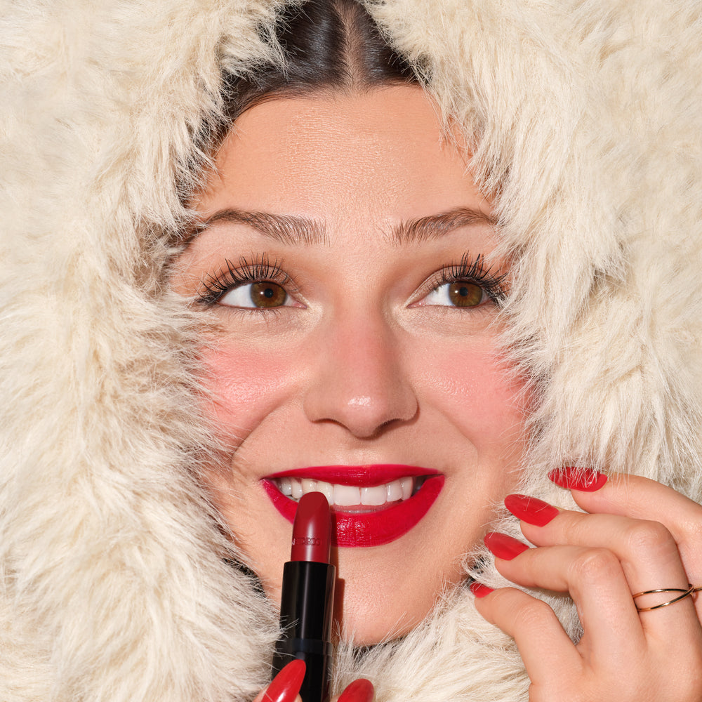 Nahaufnahme einer lächelnden Frau, deren Gesicht von flauschigem, cremefarbenem Kunstfell umrahmt wird. Sie trägt leuchtend roten Lippenstift und rote Nägel und hält einen roten Lippenstift an ihre Lippen.