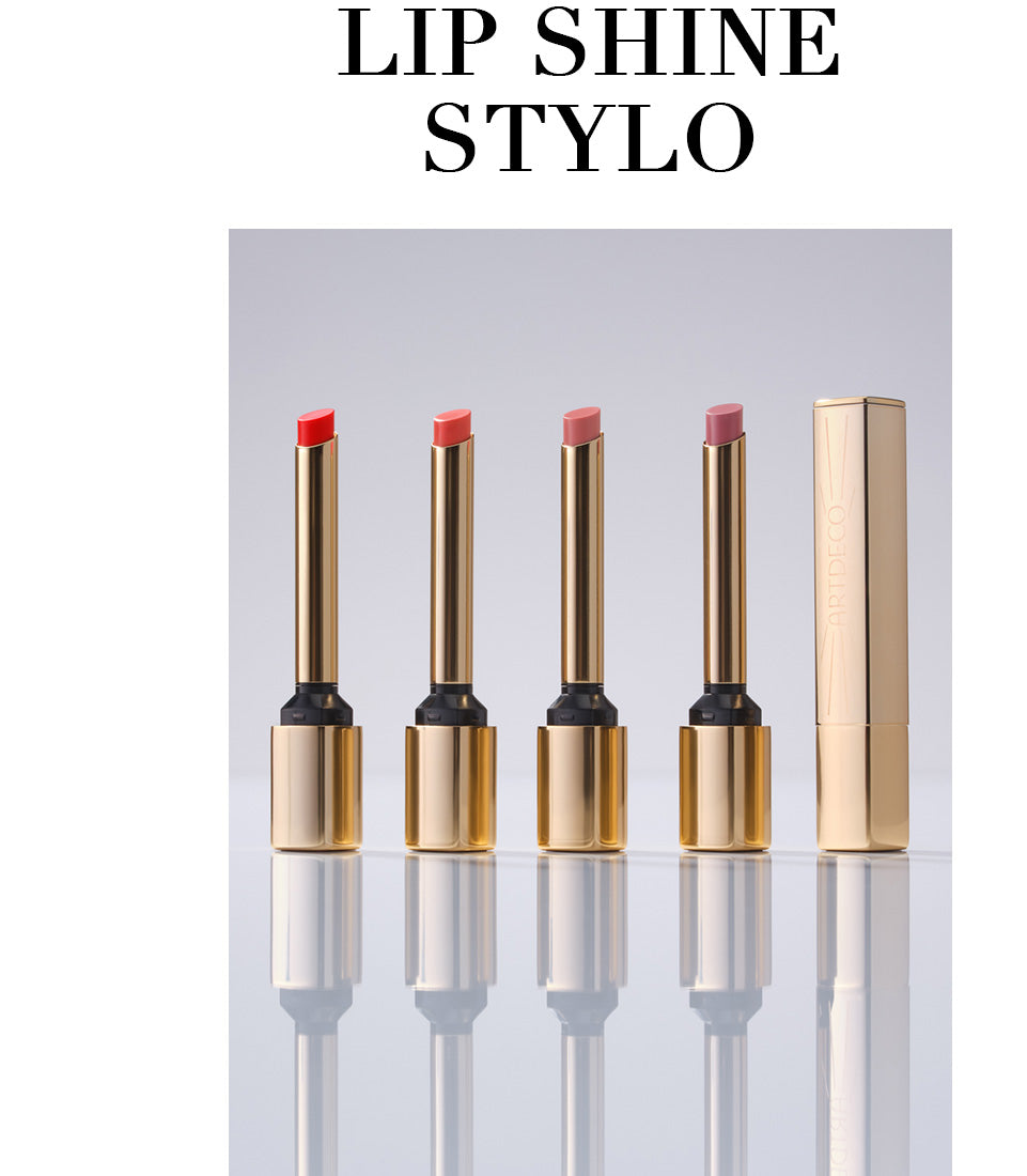 Alle vier Lip Shine Stylo stehen geöffnet aufgereiht auf einem neutralen Hintergrund. Ein fünfter Lip Shine Stylo steht geschlossen daneben. Die filigrane, goldene Verpackung vermittelt Hochwertigkeit des Produktes. Zudem steht in Großbuchstaben der Name des Produktes »Lip Shine Stylo«.