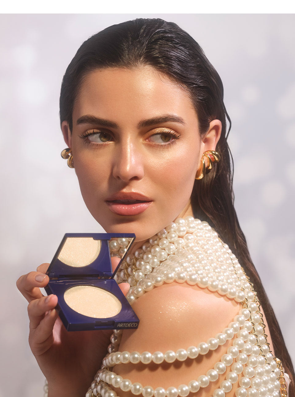 Brünettes Model blickt über ihre Schulter, hält das halbgeöffnete Diamond Beauty Powder in ihrer Hand. Der champagnerfarbene All-over-Highlight würde dabei in ihrem Gesicht als auch auf den Schultern, für einen unwiderstehlichen Glow aufgetragen