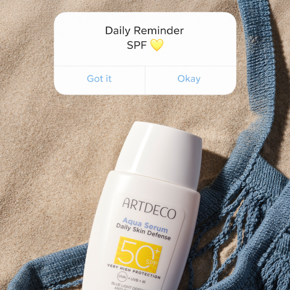 Aqua Serum Daily Defense SPF50+ liegt im Sand. Darüber wird grafisch ein Reminder zum Sonnenschutz angezeigt