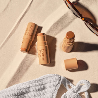 Auf Sand platzierter Bikini, Brille und Bronzer Sticks in drei Farben.