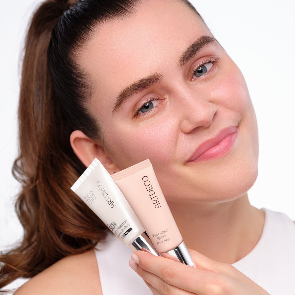 Lächelnde Frau mit Zopf hält das High Protection Hyaluronic Fluid SPF 50 und den Wonder Skin Primer am Gesicht.