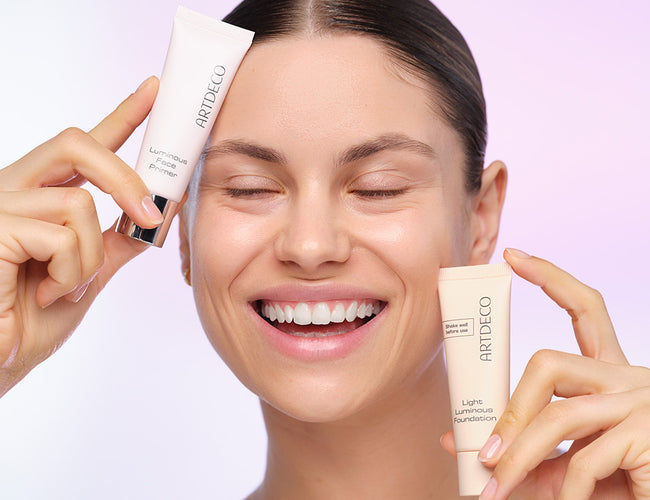 Model hält Foundation und Primer für einen natürlichen Glow beim Tages-Make-up in jeweils einer Hand und lächelt dabei 