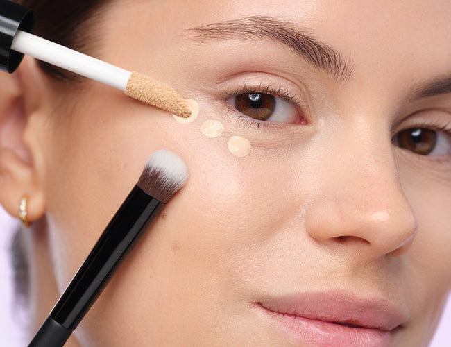 Close-up auf das Auge des Models, an welchem der Concealer punktuell aufgetragen wurde und der Applikator und Pinsel zusätzlich gezeigt wird 
