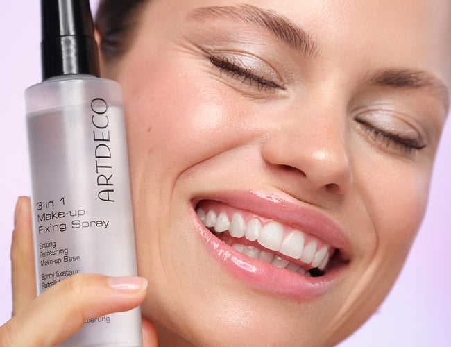 Model hält das Fixing Spray an ihre Wange, hat dabei die Augen geschlossen und lächelt breit.